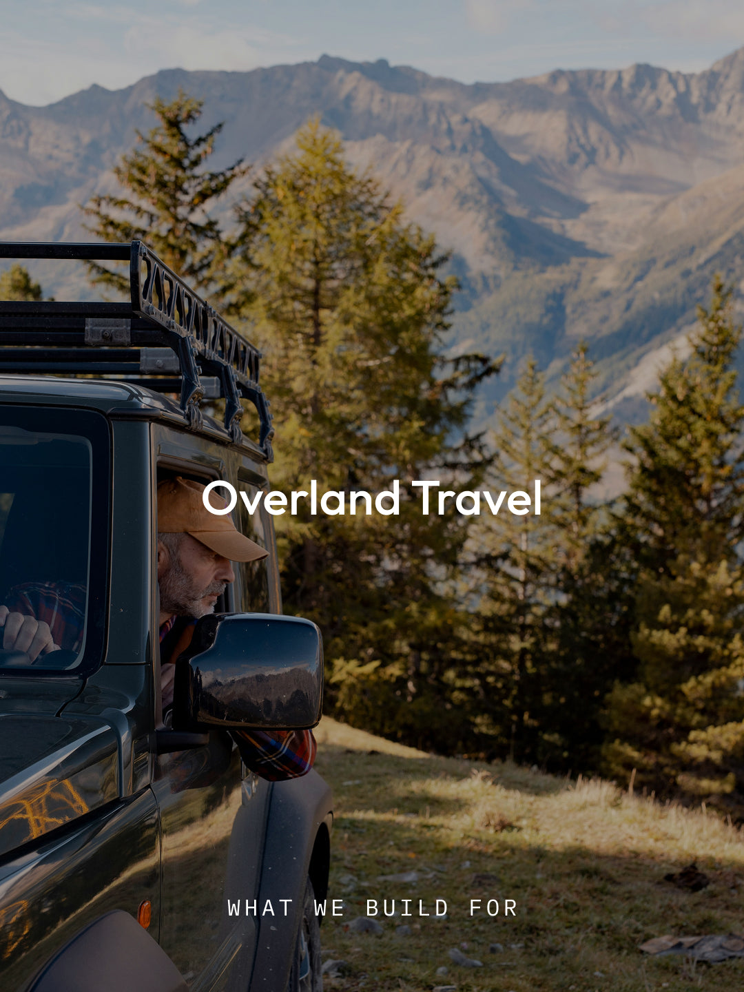A dark off-road vehicle with a roof rack is parked outdoors on a grassy hillside with mountains and evergreen trees in the background. A person is visible in the driver's seat.