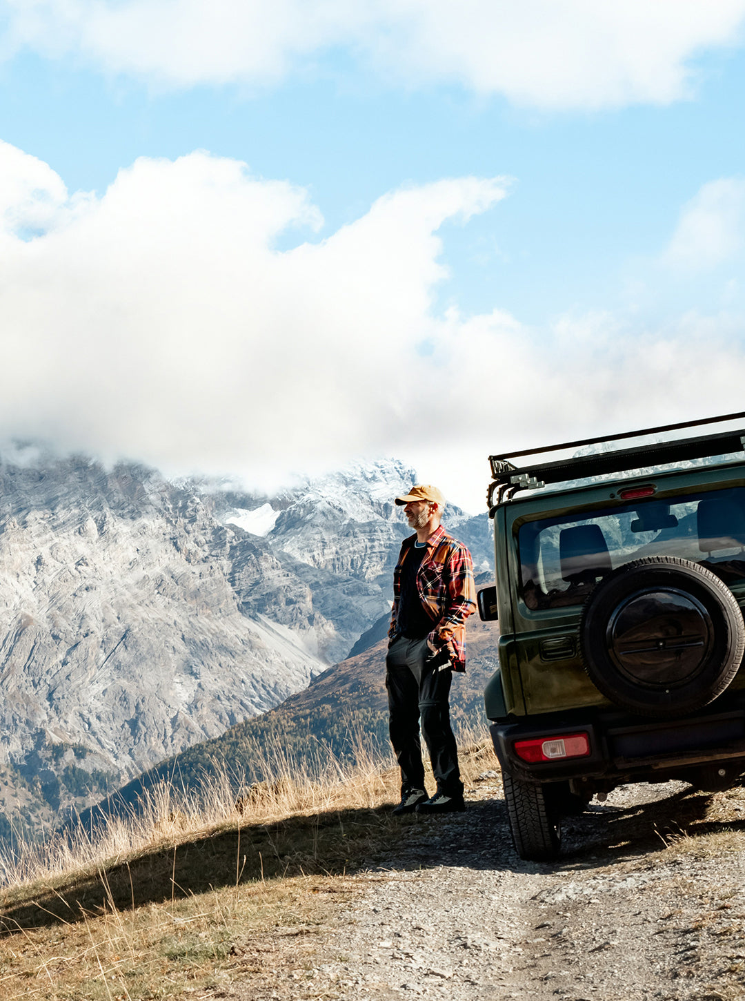 A man wearing a tan hat and a red-and-black plaid flannel shirt stands on a dirt road next to the rear of an olive green off-road vehicle, with a dramatic mountain landscape and cloudy sky in the background.