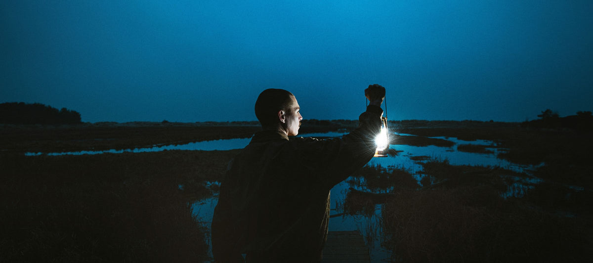 A man standing in a dark marshland at night holding a bright lantern or flashlight, with a deep blue sky and water reflections in the background.