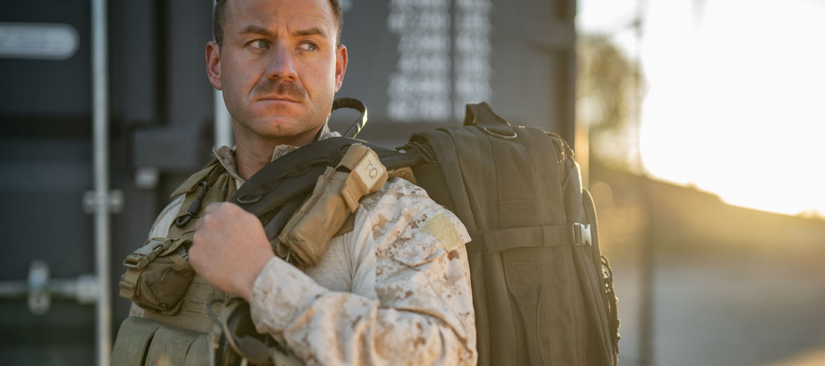 A man in military-style camouflage clothing and a large dark tactical backpack, standing outdoors with warm sunlight and a blurred background.