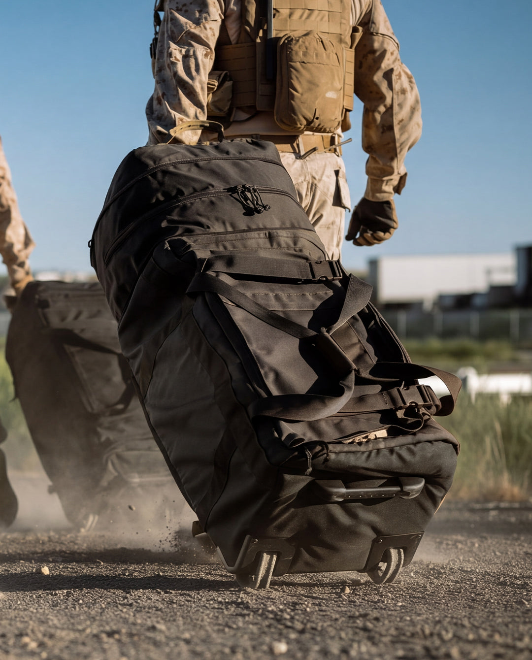 A large black tactical rolling duffel bag with wheels is being pulled by a person in desert camouflage military gear outdoors on a dusty gravel path.